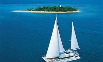  A large catamaran style boat with sales in the middle of the ocean with a tropical island in the background