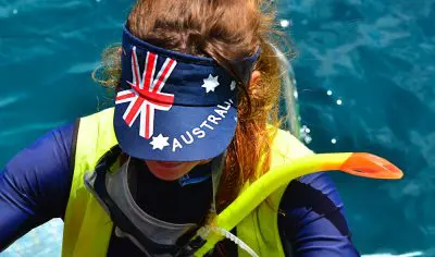 Traveller in an Australia visor, snorkel, life vest, and rucksack stands by vibrant blue ocean water under a clear sunny sky.