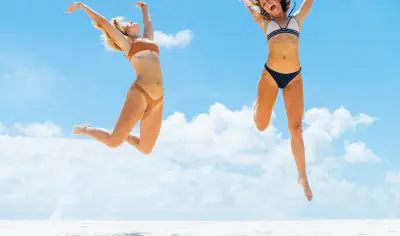 Two women in colourful bikinis jump with excitement on a sunny beach, highlighting unforgettable moments on a 16-day budget travel package.