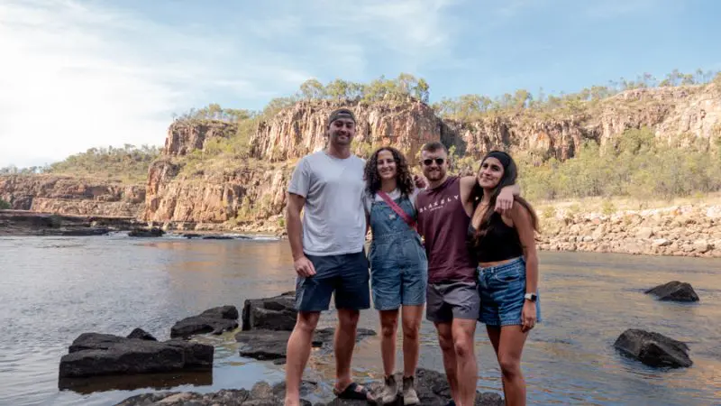 Four friends smiling on river rocks during a 3-Day Kakadu Adventure Camping tour with scenic cliffs and lush trees in the background.
