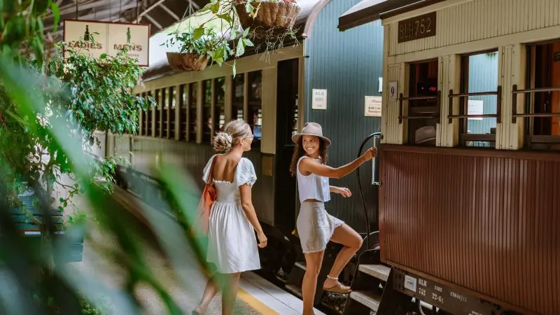Two women in summer outfits board a vintage train at a leafy station, ready to journey on the scenic skyrail.