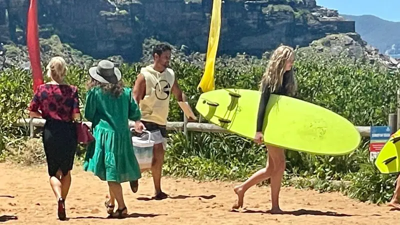 Four people walk along a sandy path, filming possibly for Celebtime Tours or 1 Day Home Away Tour, with a bright yellow surfboard nearby.
