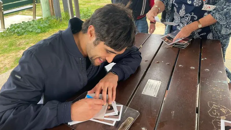 A celebrity signs autographs at a Celebtime Meet an Actor tour, sitting at a picnic table whilst fans eagerly wait in queue nearby.