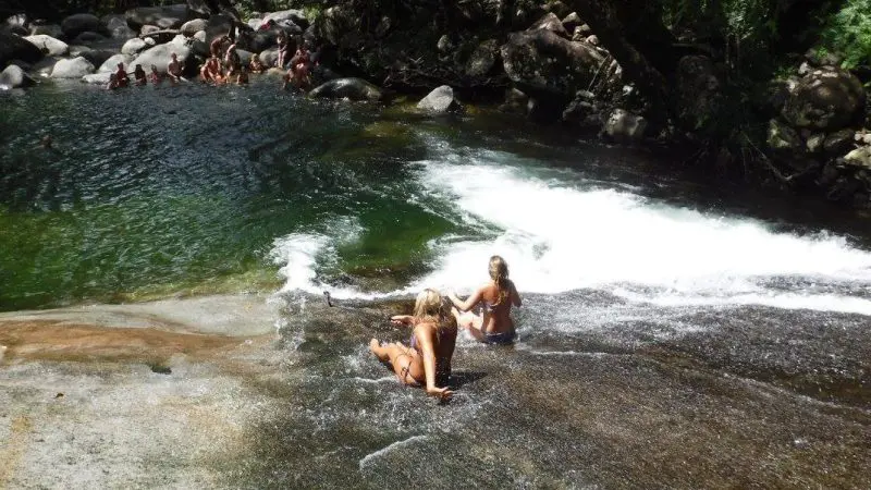 Two people relaxing on a wet rock beside a stunning waterfall during a 1 Day Rainforest Tour with Waterfall Wanderers adventure.