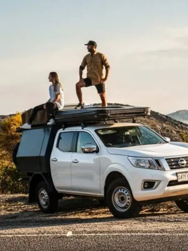 Couple stands atop a white pickup lorry overlooking a stunning coastal landscape with rolling hills and blue sea in the background.