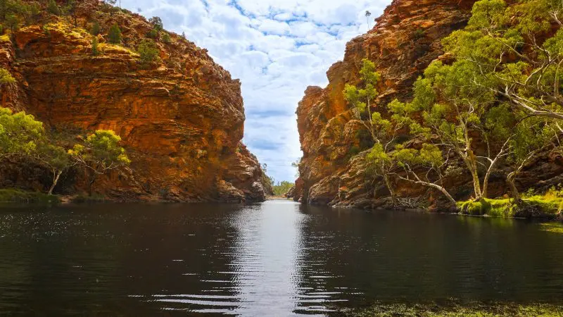 Scenic narrow gorge with towering red rock cliffs, tranquil river, lush green trees, and a partly cloudy sky—stunning nature view.