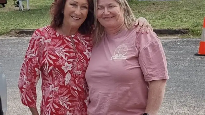 Two smiling women in red floral and pink tops by a serene lake during Celebtime Tours 1 Day Home Away Tour, enjoying scenic views.