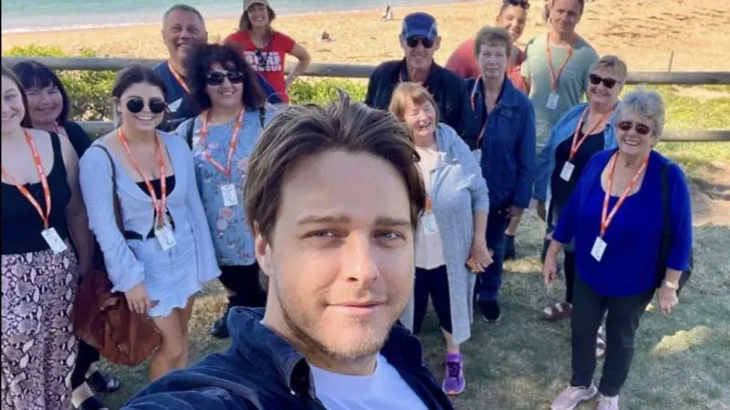 Group enjoying a 1 Day Home Away Location Tour, smiling for a beach selfie with crystal blue ocean waves in the scenic background.