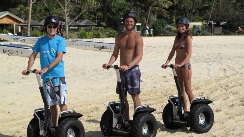Three adventurers in summer attire ride Segways along a scenic sandy beach on the Adventure Moreton Island Wrecks Adventure Tour.
