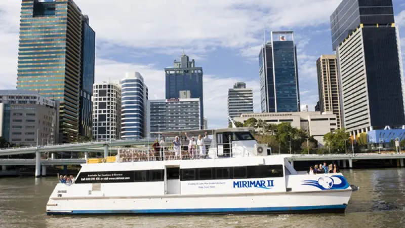 Scenic white river cruise boat with passengers sails by Brisbane city skyline, offering return trips to Lone Pine Koala Sanctuary.