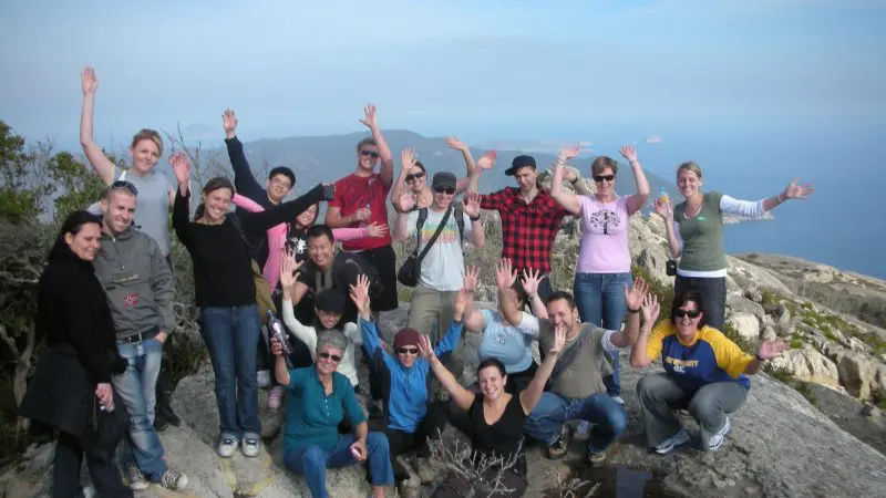 Smiling tour group on rocky Wilsons Promontory hilltop with ocean backdrop, enjoying 1 Day Go West Tours adventure experience.