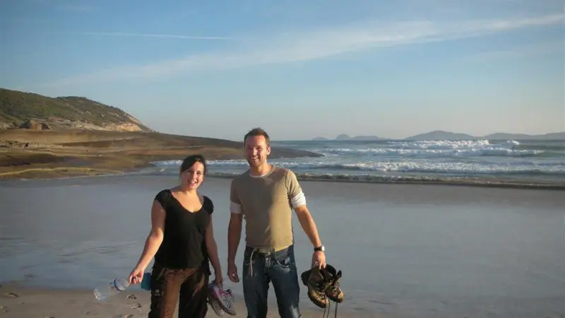Two happy travellers stand barefoot on Wilsons Promontory’s sandy beach with Go West Tours, grinning beside scenic ocean waves.