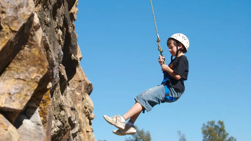 Child wearing helmet abseils down outdoor climbing wall with instructor, surrounded by blue sky and trees, enjoying adventure.