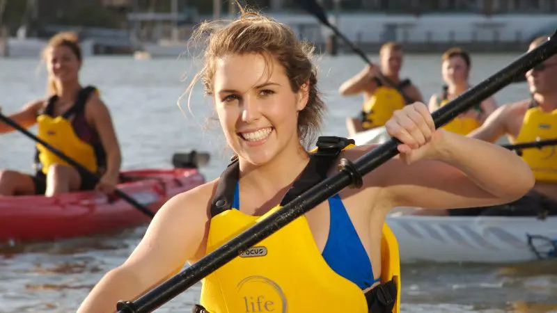 Smiling woman kayaking on a river during a 1–5 hour kayak hire, with others in yellow life vests paddling behind her for adventure.