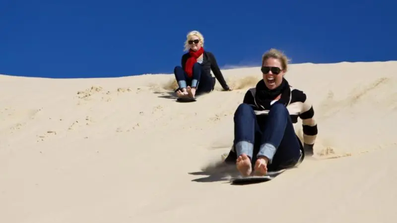 Two women sandboarding on golden dunes under the sun, smiling during a 1 Day Whale Watching Port Stephens adventure tour.