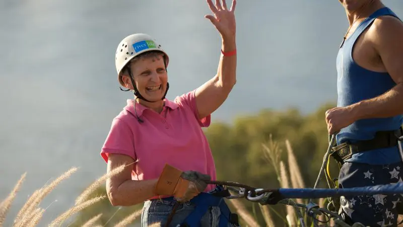 Elderly woman smiles and waves, wearing a helmet while ziplining outdoors with a certified rock climbing guide by her side.