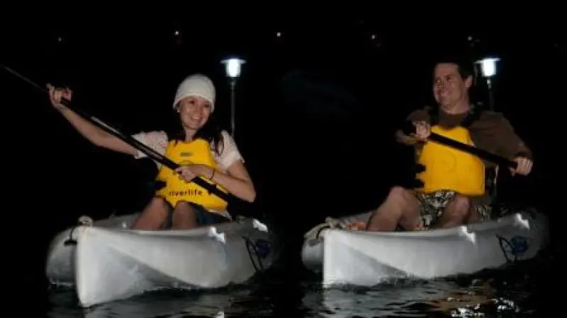 Two people in life jackets kayak at night with vibrant lights on their craft, enjoying a scenic Friday outing; catering not provided.