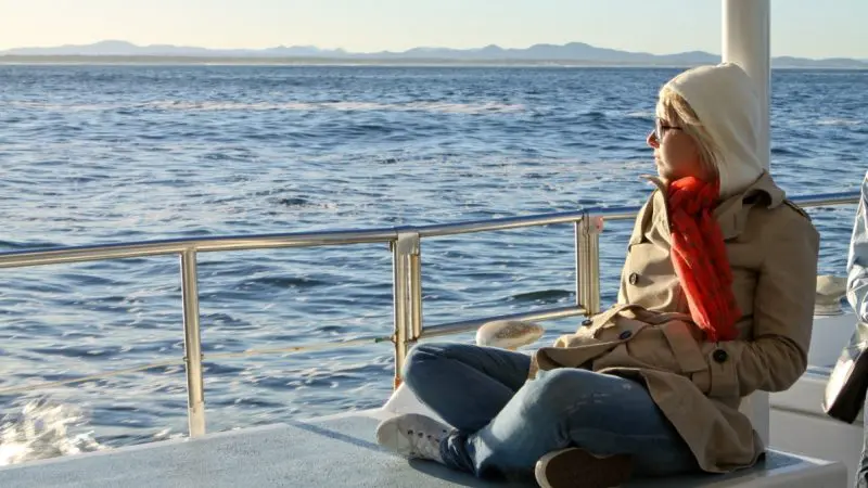 Person in coat and red scarf aboard 1 Day Whale Watching Port Stephens boat, looking at scenic mountains under clear blue sky.
