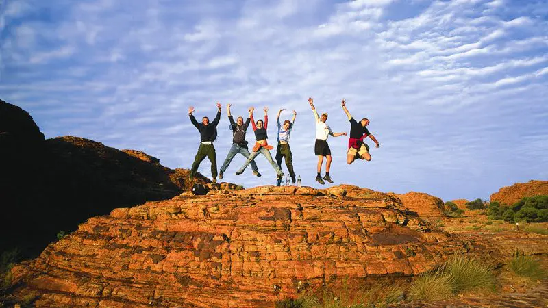 A group of six friends leap on a scenic rocky outcrop with vibrant blue skies during a 3-night Uluru adventure from Alice Springs.