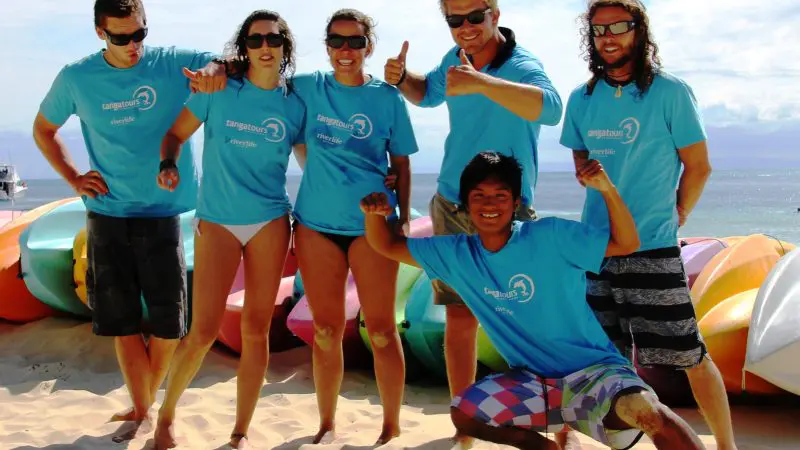 Group of five in matching blue shirts smiling on Moreton Island’s sandy beach, colourful kayaks nearby, Adventure Wrecks Tour fun.