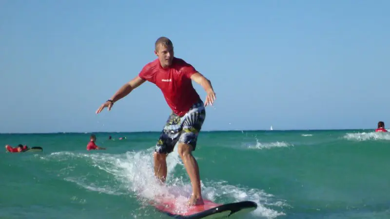 Man in bright red shirt skilfully surfs a small wave at Learn To Surf Australia's Longest Wave, clear blue sky overhead.