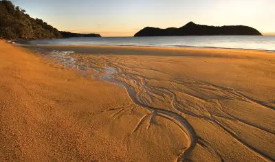 Breathtaking golden sandy beach at sunset, Three Day Classic route, with sparkling water streams and rolling hills by a tranquil sea.