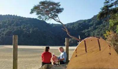 Two campers relax by a bright yellow tent on a Three Day Classic, nestled among lush forested hills and trees beneath a sunny, clear sky.