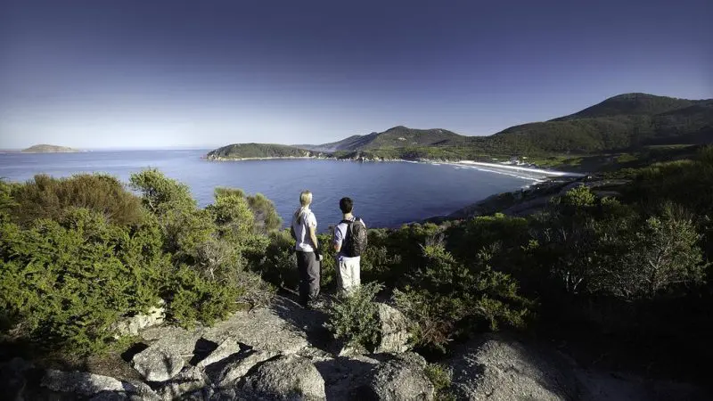 Two people stand on rugged rocks, taking in panoramic views of Wilsons Promontory Bay during a Go West Tours 1 Day guided tour.