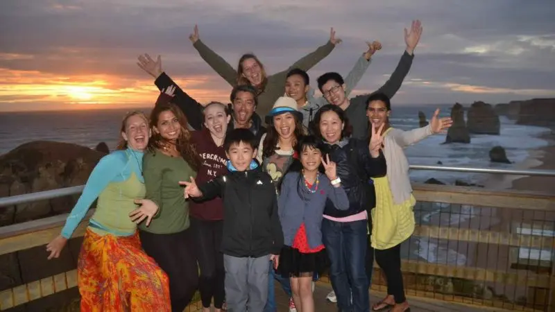Joyful group with raised arms poses on a scenic boardwalk at sunset during Go West Tours’ 1 Day Great Ocean Road Sunset Tour.