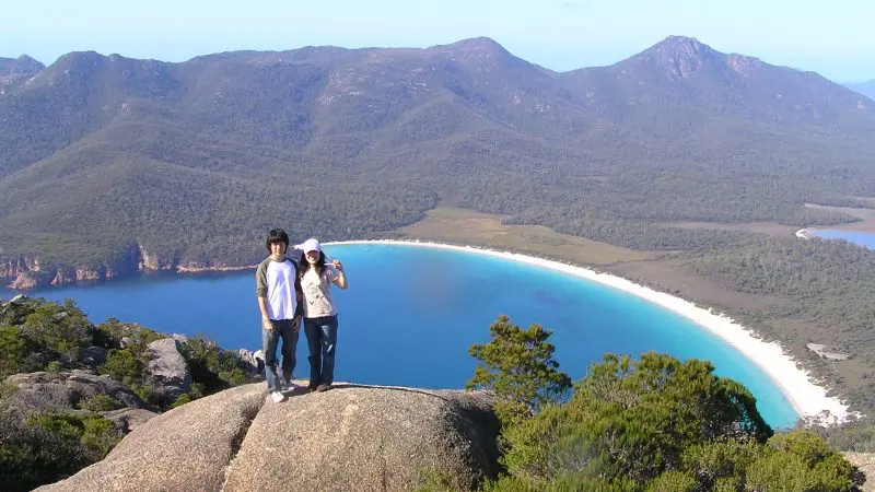 Two people atop a large rock admire stunning bay views, pristine white sand, and lush forests on the 6 Day Tasmania Explorer Tour.