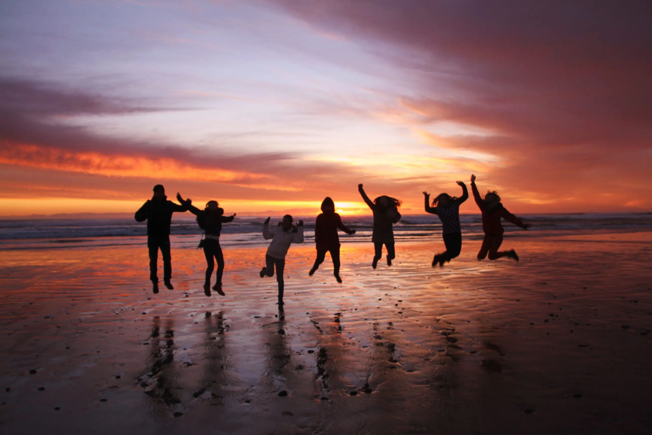 Seven people leaping with excitement on a beach at sunset during the 5 Day Famous 5 Tasmania Tour, showcasing travel joy and adventure.