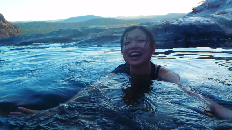 Smiling woman swimming in crystal-clear Kakadu waters at sunset, framed by hills and rock formations—a top-rated tour highlight.