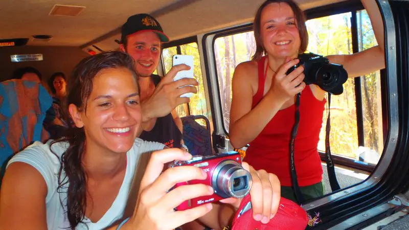 Three tourists smiling on a Kakadu National Park tour capture photos from their safari vehicle, enjoying a sunny day before heading to their hotel.