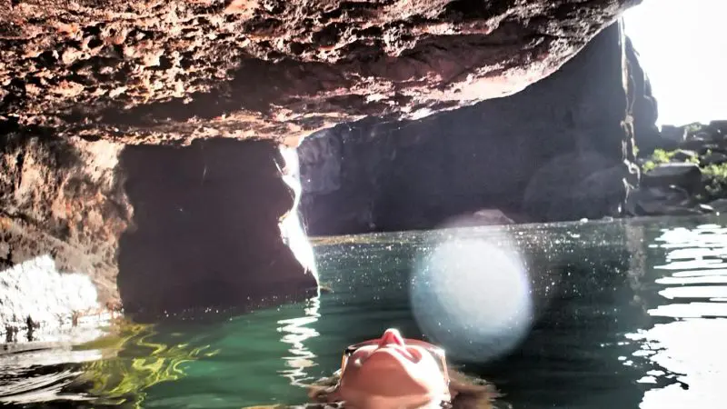A person in a vibrant red bikini floats in crystal-clear water beside a dramatic rocky overhang, resembling top Kakadu attractions.