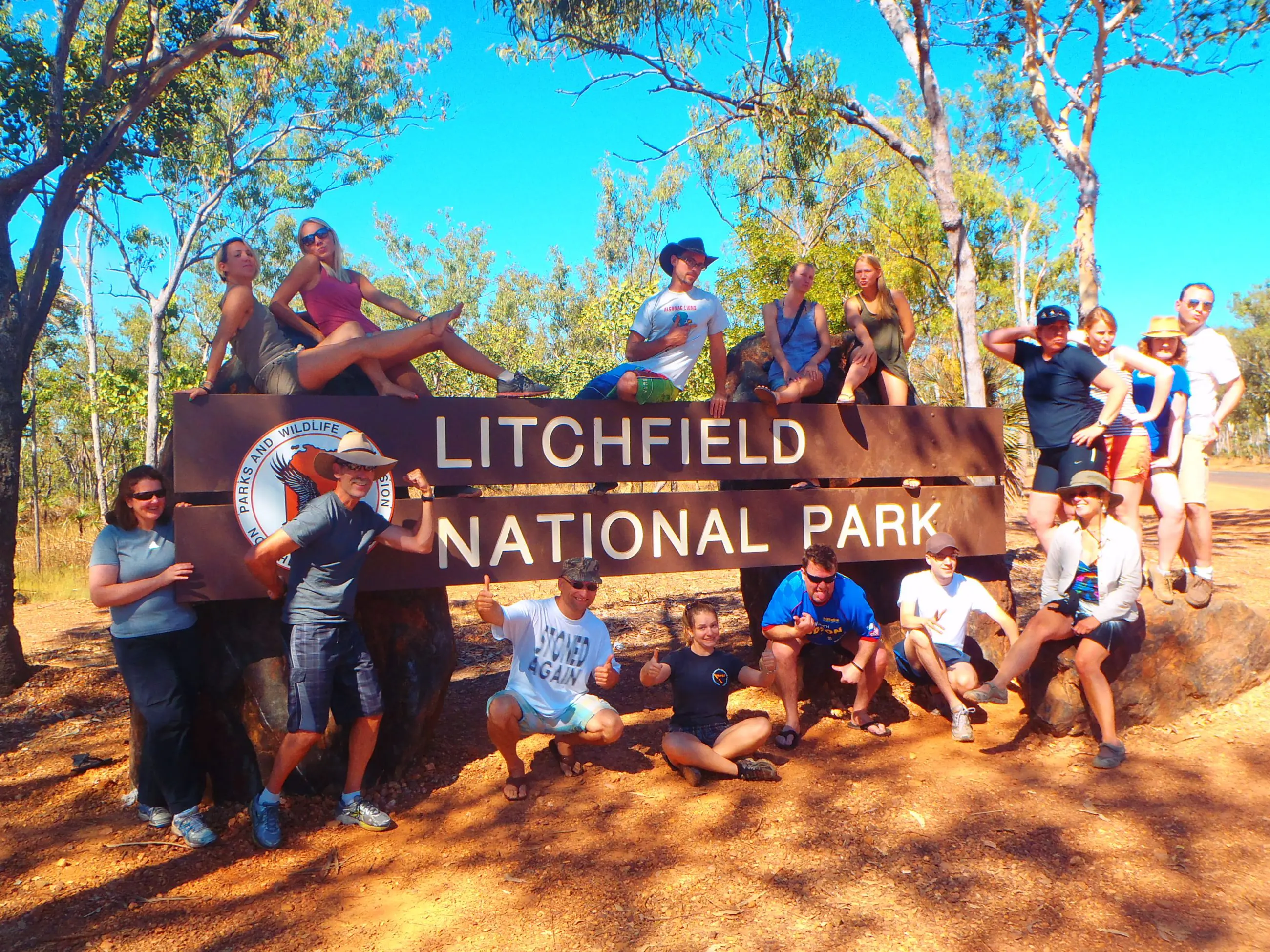 Tour group enjoying a 3-Day 4WD Kakadu Litchfield adventure, posing by the national park sign under sunny skies in lush woodland.