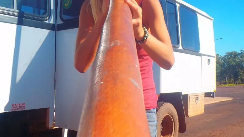 A woman wearing sunglasses plays a didgeridoo outdoors by a branded Kakadu Tour vehicle under bright sunshine in Australia.