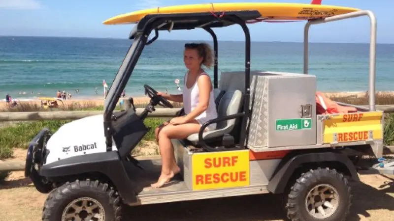 Woman enjoying a sunny Celebtime 1 Day Home Away Location Tour, seated in a surf rescue vehicle with yellow surfboard by the beach.