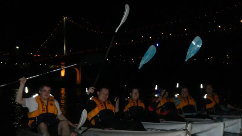 Six kayakers in life jackets paddle at night, illuminated by city lights and a glowing bridge in the background on a Friday evening.