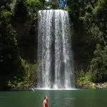 A woman stands by Paronella Park’s scenic waterfall amid vibrant rainforest, whilst another person swims in the refreshing natural pool.