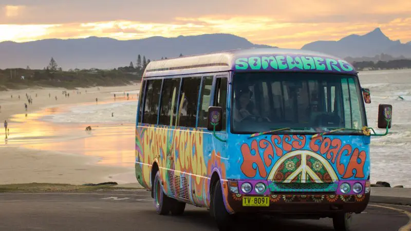 Vibrant, hippy-style Happy Coach Byron Bay bus parked on a scenic beach at sunset with mountains in the background.