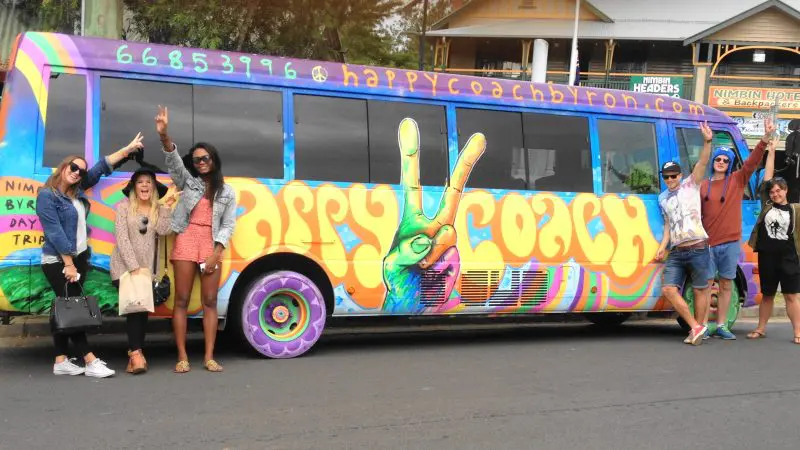 Six happy travellers pose by the vibrant Happy Coach bus during a 1 Day Nimbin Tour from Byron Bay, ready for their adventure.