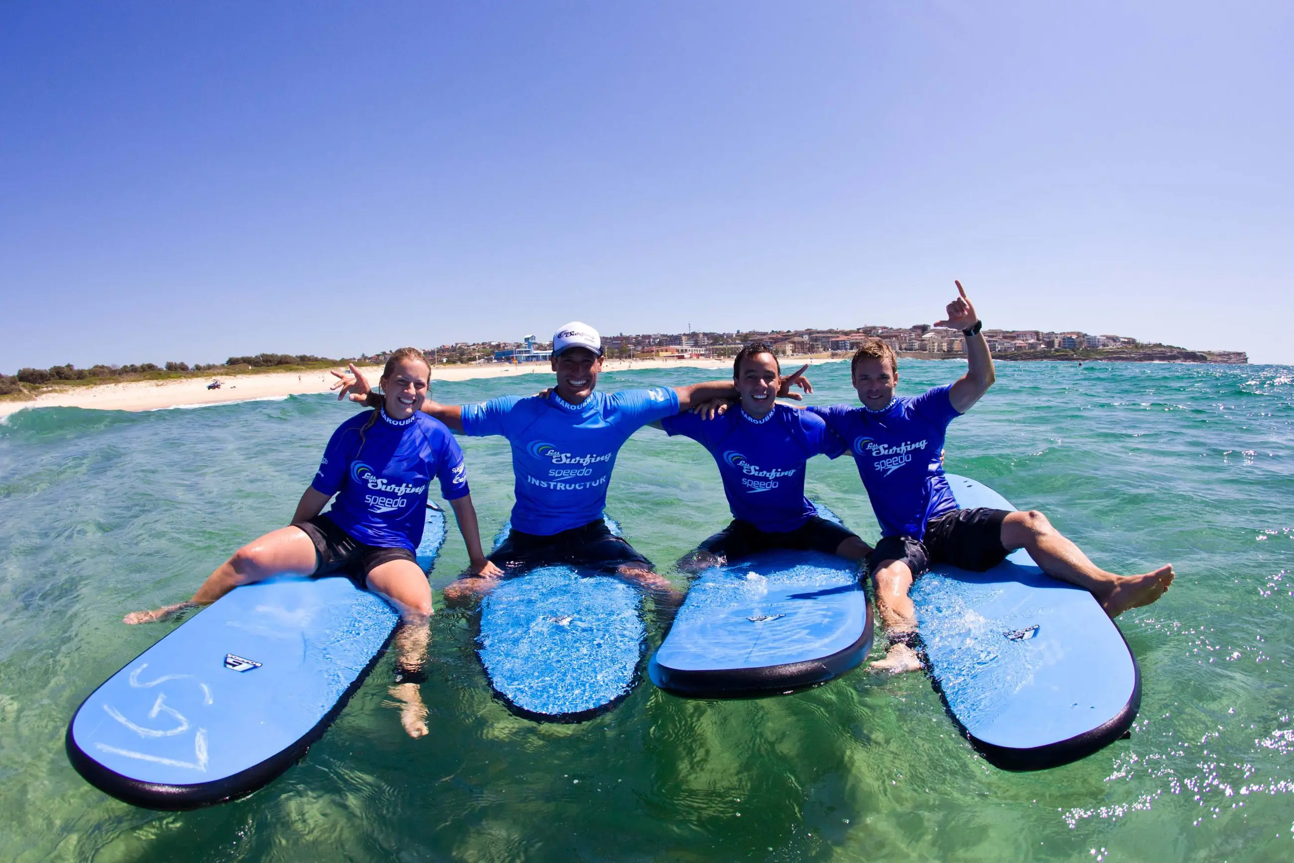 Smiling group in matching blue shirts pose confidently on surfboards under sunny skies during a Byron Bay Learn to Surf Half Day Tour.