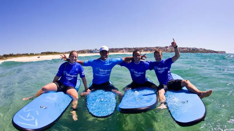 Four happy surfers in blue shirts sit on boards at a Byron Bay 2 Hour Small Group Surf Lesson, smiling brightly under a sunny sky.