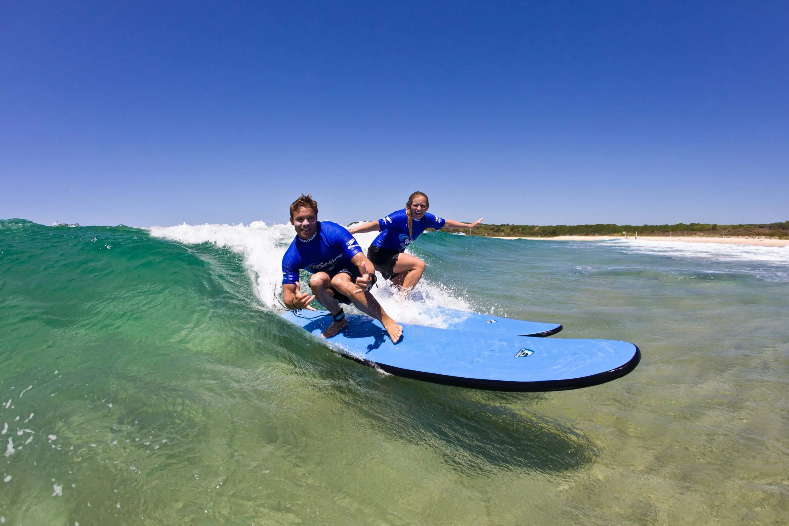 Two surfers in blue shirts ride gentle waves at Byron Bay during a two-hour small group surf lesson under clear, bright blue skies.