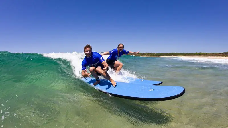Two surfers in blue shirts ride gentle waves at Byron Bay during a two-hour small group surf lesson under clear, bright blue skies.