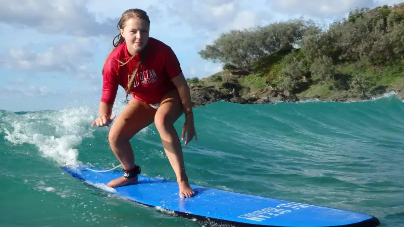 Young woman in red shirt surfing near coastal rocks at Australia’s Longest Wave Beach Drive by Learn To Surf—expert surf lessons.