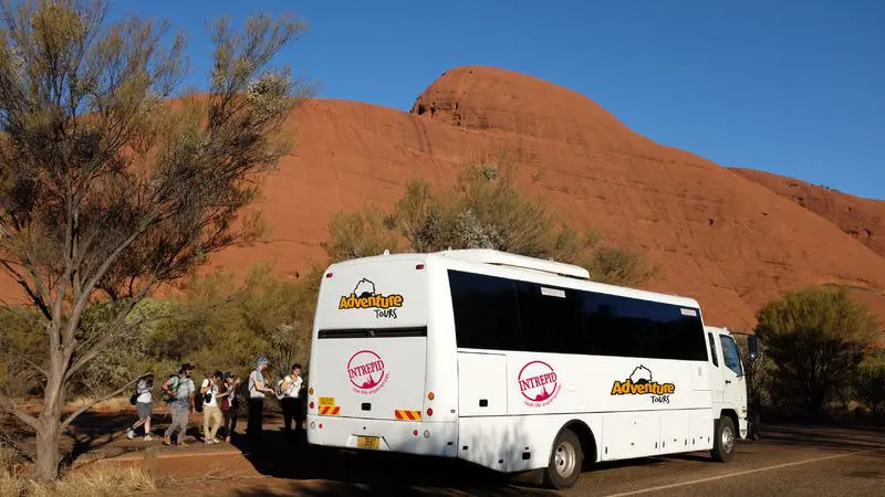 A white tour coach for the 3 Day Uluru Rock The Centre Tour is parked by iconic red Uluru rocks as tourists explore on foot nearby.