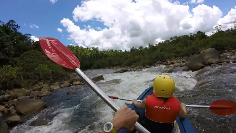 Adventurous duo kayaking on a rugged river at Mission Beach, safety gear on, navigating rapids beneath partly cloudy skies.