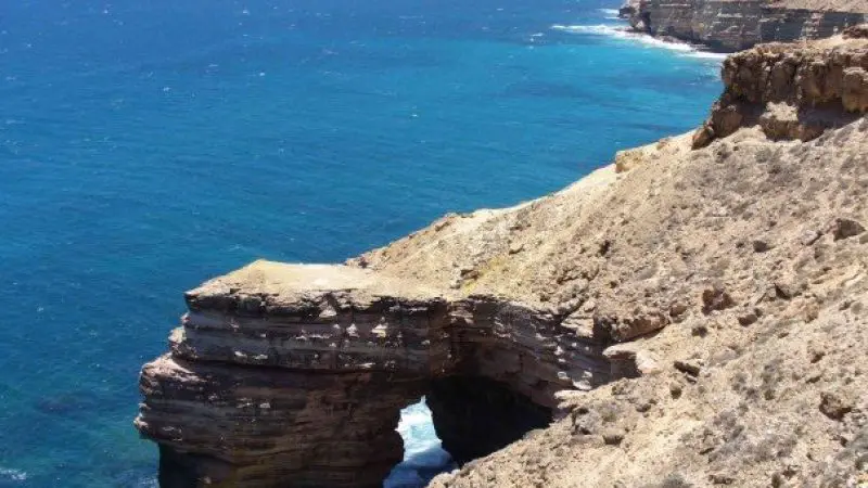 Dramatic rocky coastal cliffs and a natural stone arch over turquoise ocean waves, featured on the 6 Day Perth to Exmouth Tour.