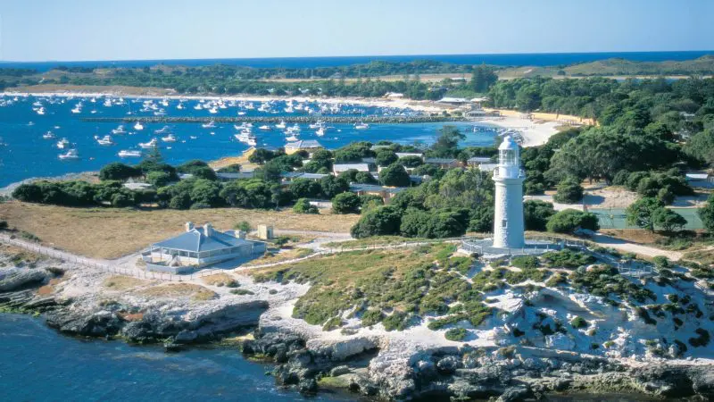 Iconic white lighthouse on Rottnest Island’s rocky coast, overlooking turquoise sea and boats near Oliver Hill Train Tunnel Tour.